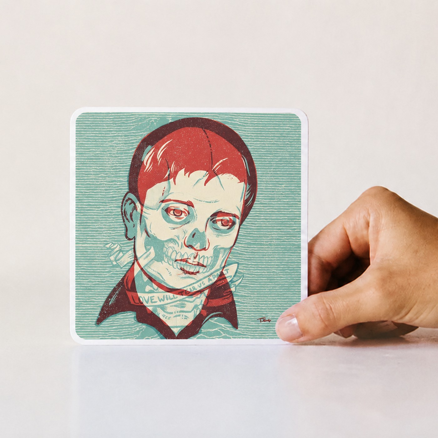 A hand holding a riso print by Travis Knight of an illustrated red-haired skull-face figure with 'Love Will Tear Us Apart' lettering